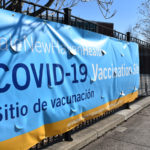 A YNHH banner showing the location of a COVID-19 vaccination site hangs on a metal fence outdoors during the daytime.
